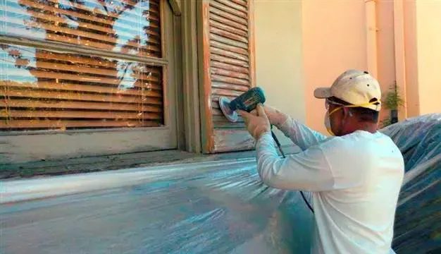 Worker in white clothing spray-paints a house exterior wall and window frame with a spray gun.