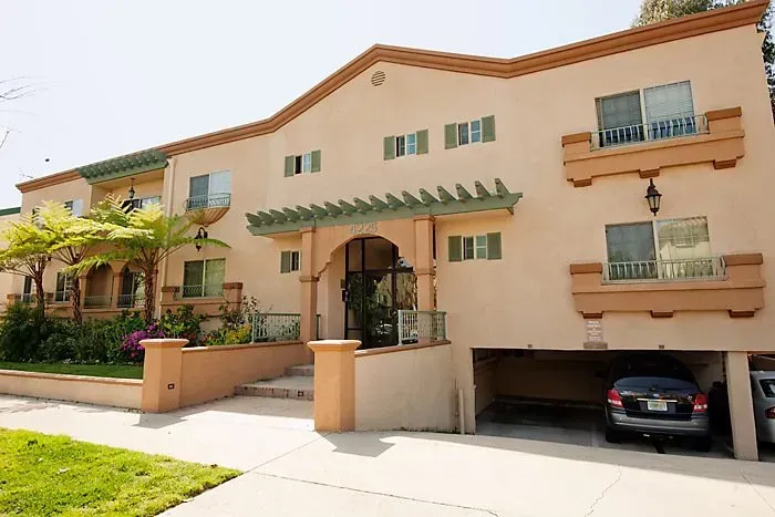 Tan stucco apartment building with green awnings, front steps, and a car parked in a driveway.
