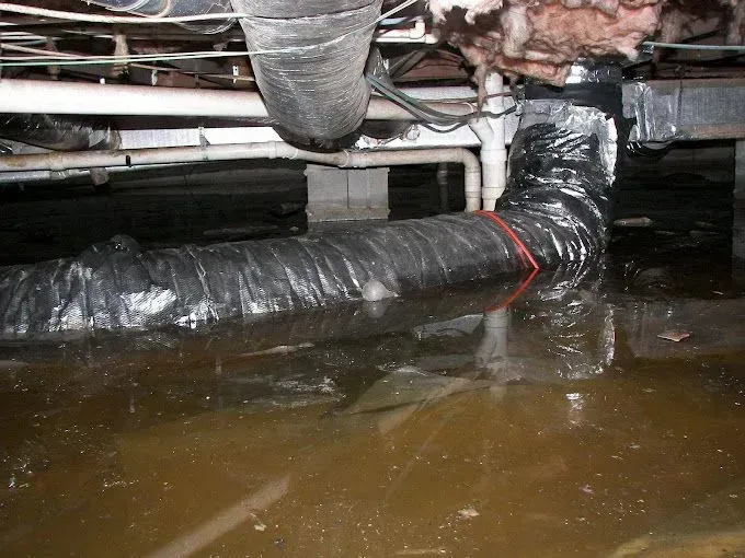 Flooded crawl space with insulated pipes and standing brown water beneath a house