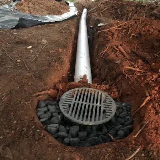 Drain pipe with round grate in a dirt trench, surrounded by gravel