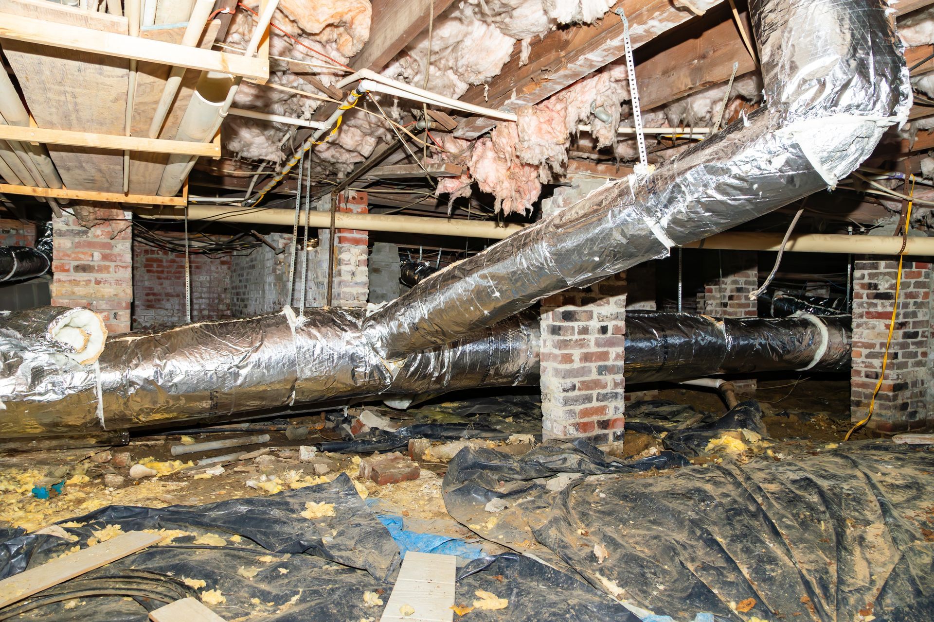 Crawlspace with exposed pipes, insulation, and damaged black floor covering under a house
