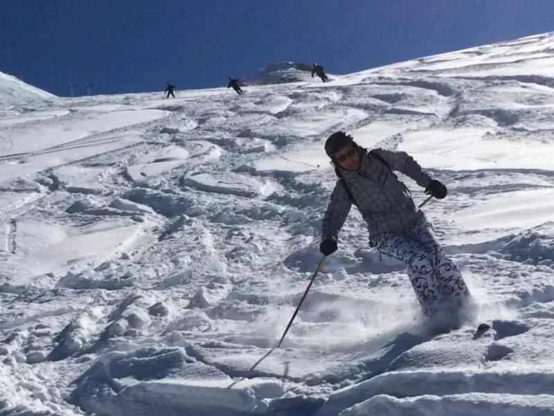 Een skiër snijdt door verse sneeuw op een zonnige berghelling, met andere skiërs op de achtergrond.