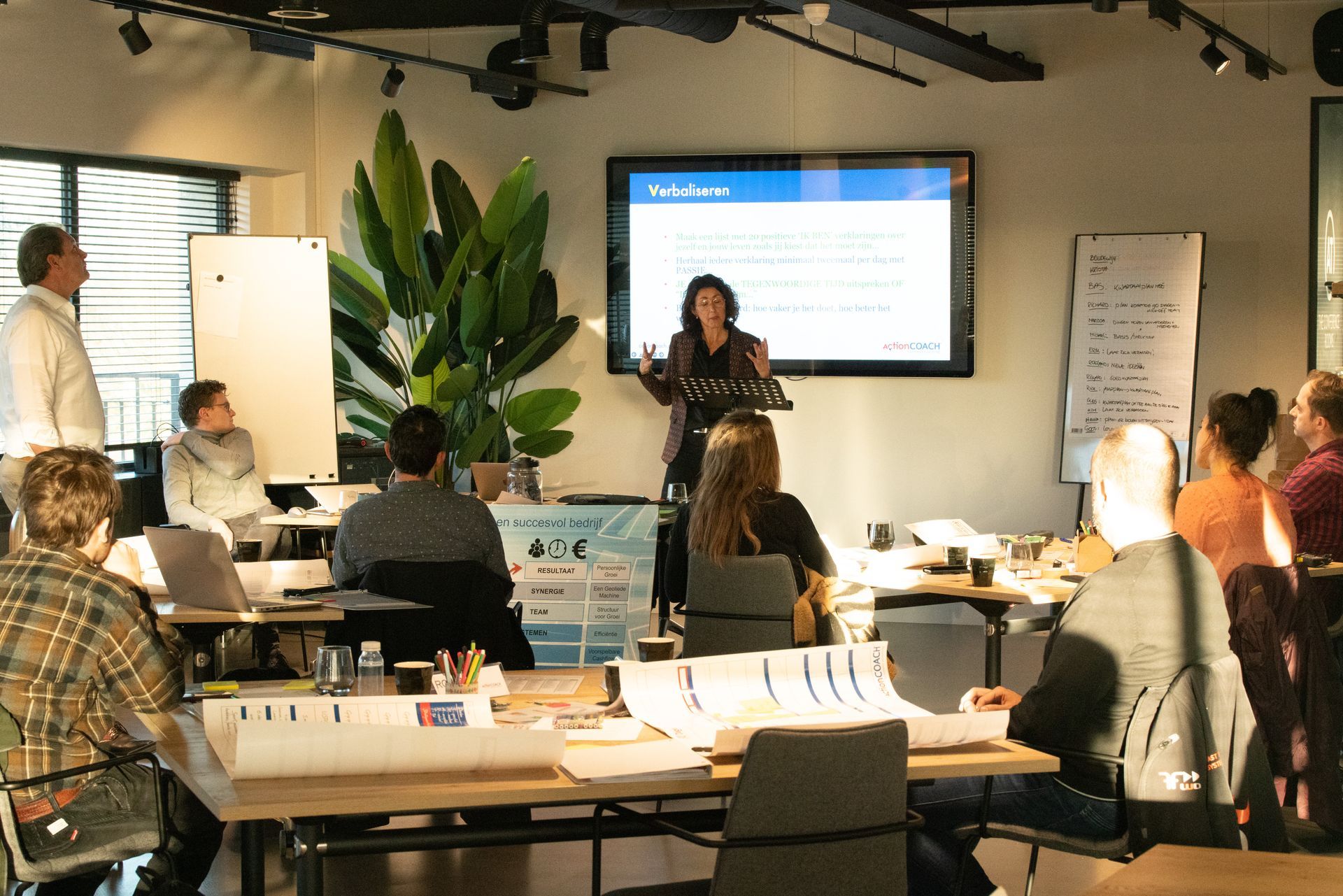 Workshoppresentatie met publiek dat rond tafels zit, spreker vooraan naast een groot scherm in een vergaderruimte.