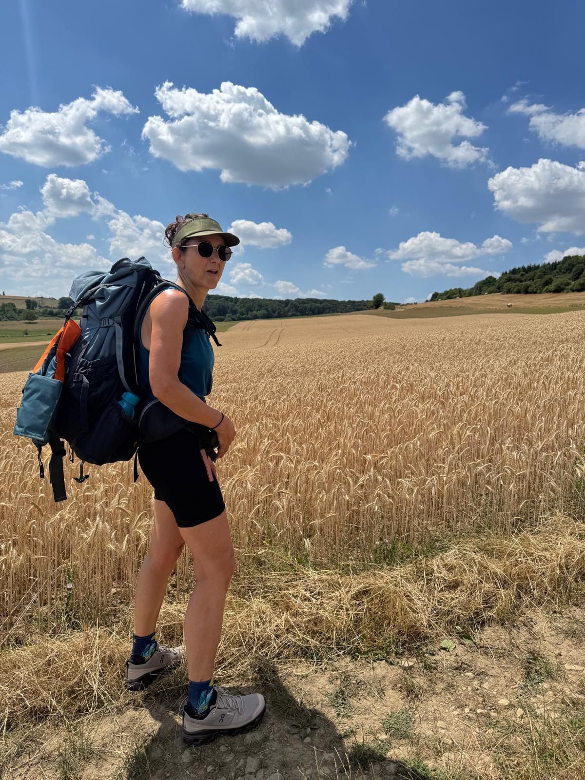 Een backpacker staat op een zandpad naast een tarweveld onder een blauwe hemel met wolken.