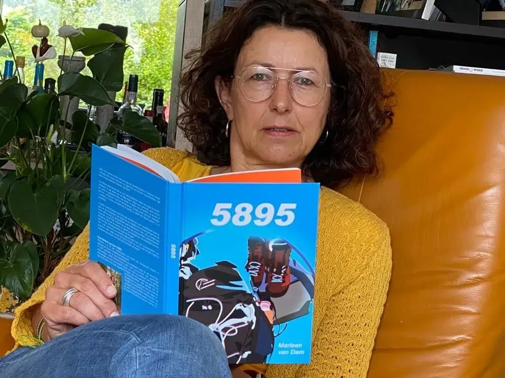 Vrouw in een gele trui leest een blauw boek binnenshuis, zittend op een oranje stoel bij een raam.