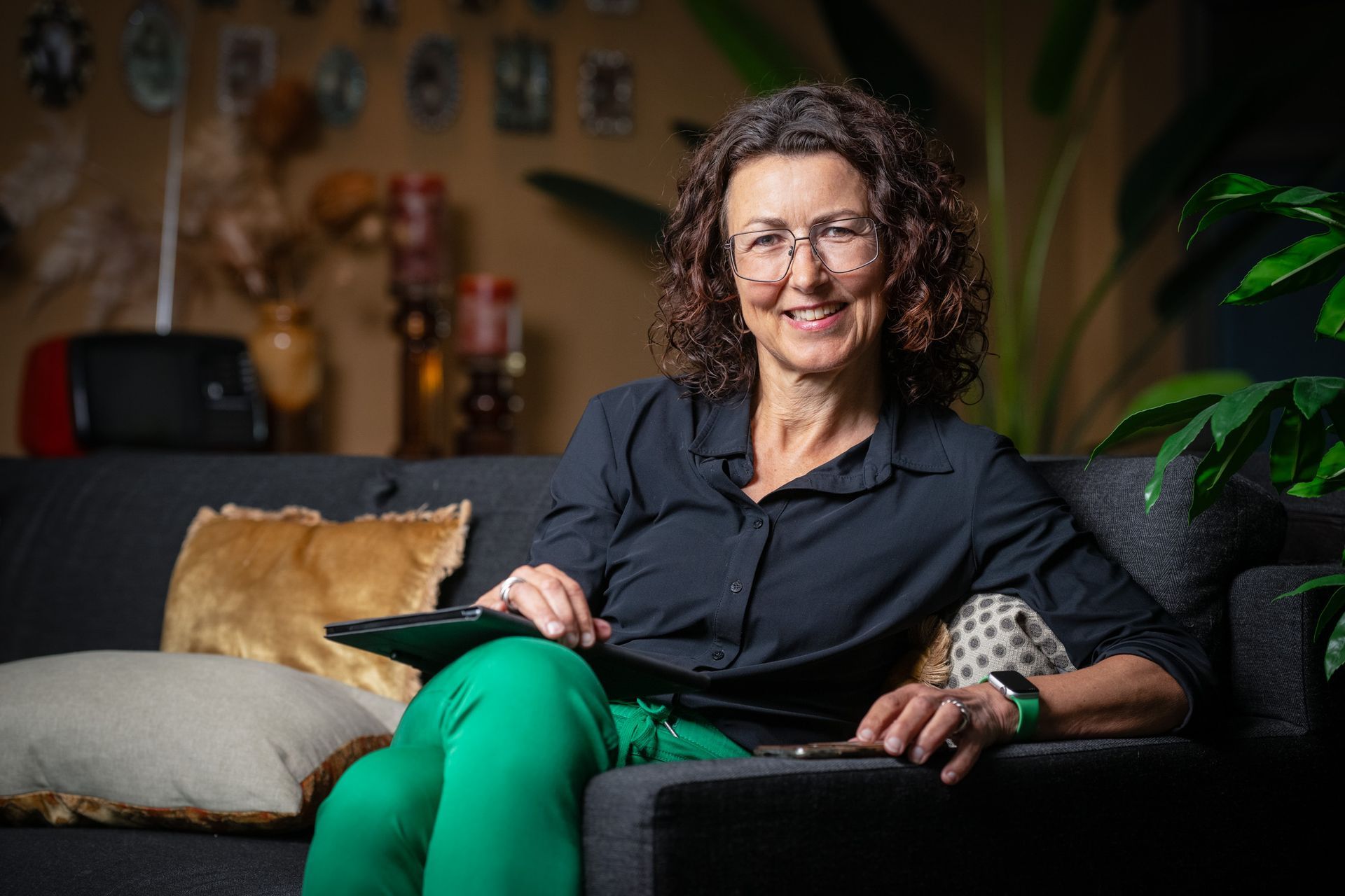 Vrouw zit met een groene broek aan op een bank en lacht in een gezellige woonkamer.