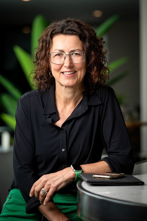 Een vrouw in een zwarte blouse glimlacht naast een tafel met een groene stoel in een vaag wazige kantooromgeving.