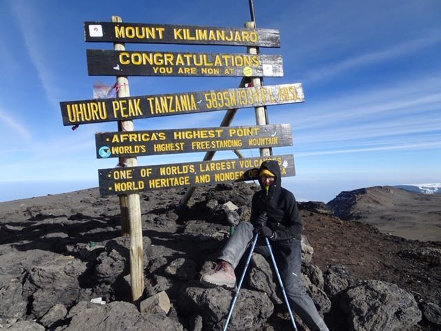 Wandelaar die uitrust naast de topborden van de Kilimanjaro op een rotsachtige bergtop onder een blauwe hemel.