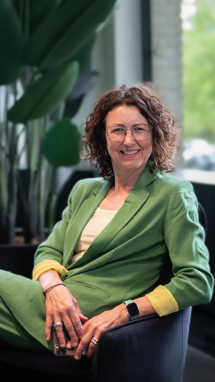 Een lachende vrouw in een groene blazer zit binnen bij een raam, met planten op de achtergrond.