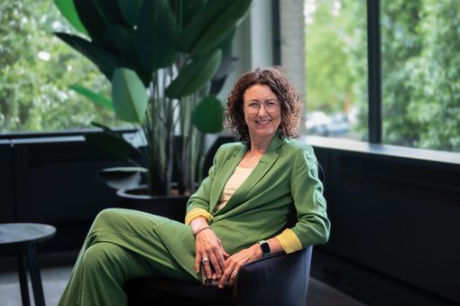 Een persoon in een groen pak zit op een stoel bij een raam met grote planten.