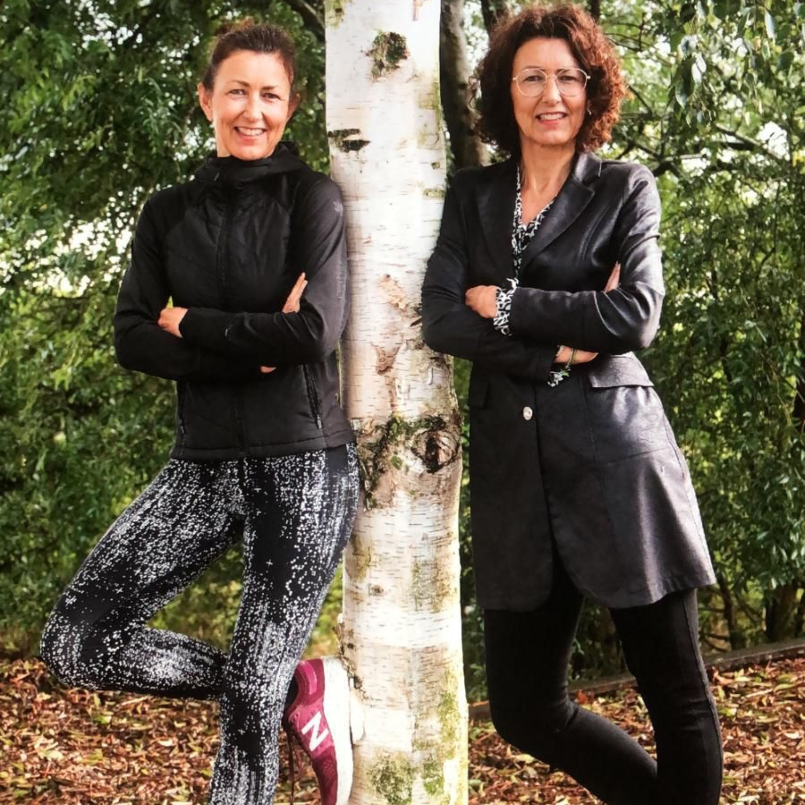 Twee vrouwen in zwarte kleding leunen tegen een berkenboom in een bosrijke omgeving.
