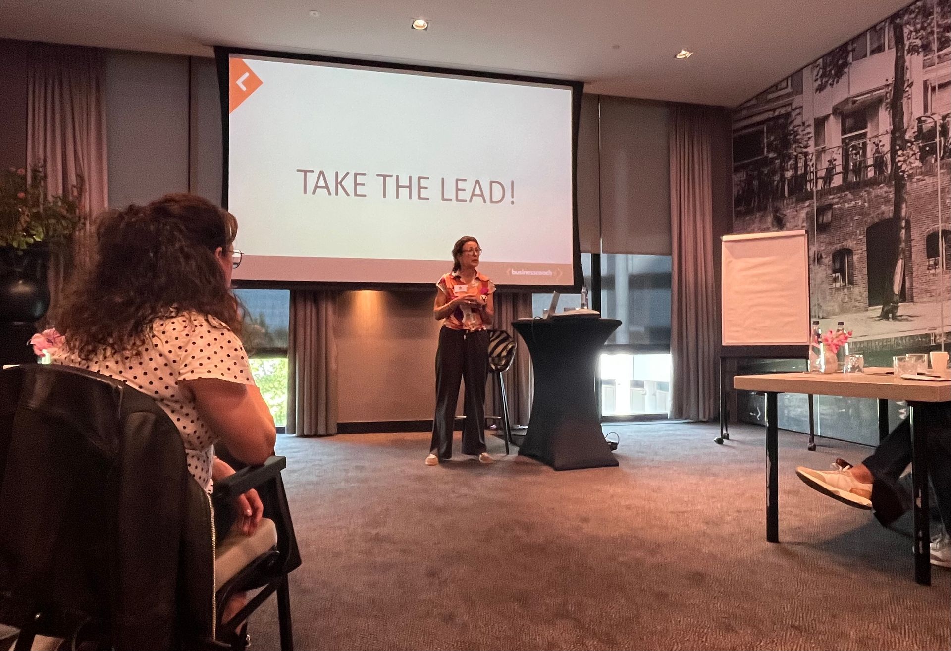 Marleen op het podium in een vergaderzaal van een hotel, met de tekst