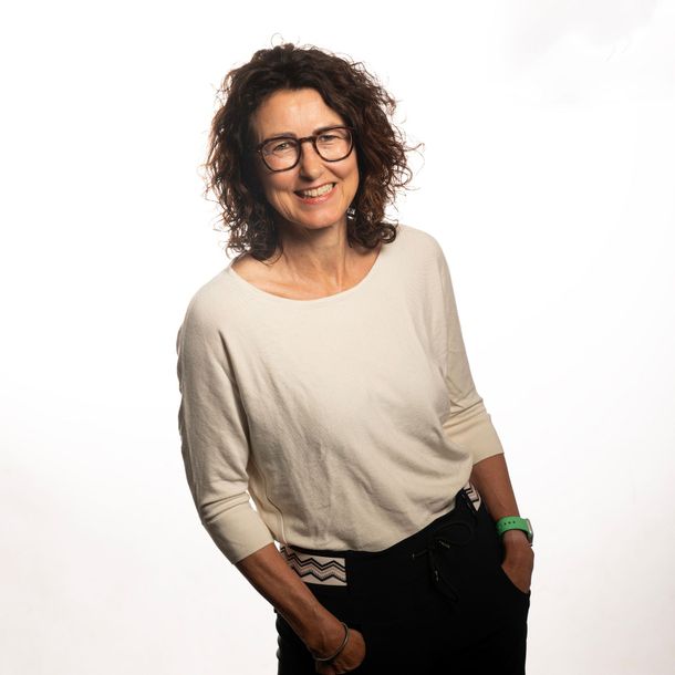 Een lachend persoon met krullend haar en een bril, gekleed in een crèmekleurig topje, tegen een witte achtergrond.