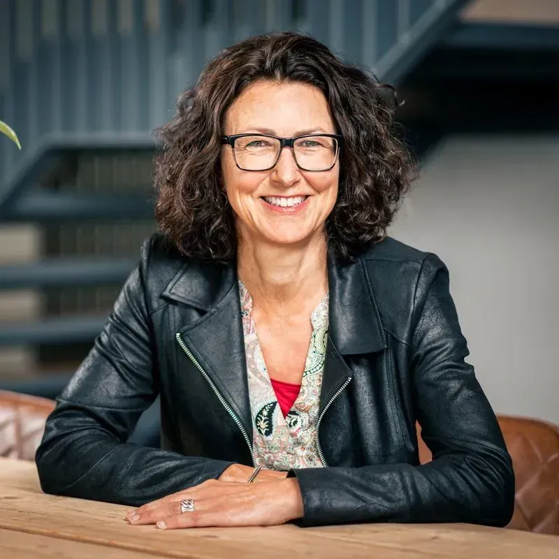 Een glimlachende persoon met een bril en een zwarte leren jas zit aan een tafel voor een trap.