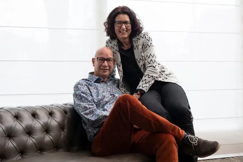 Twee mensen poseren lachend op een gecapitonneerde bank in een helderwitte kamer.