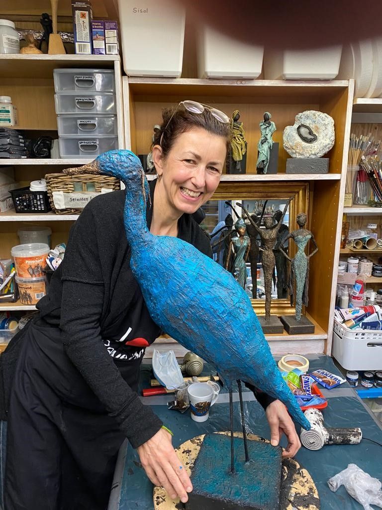 Vrouw poseert met een groot blauw vogelbeeld in een atelier, omringd door kunstwerken en planken.