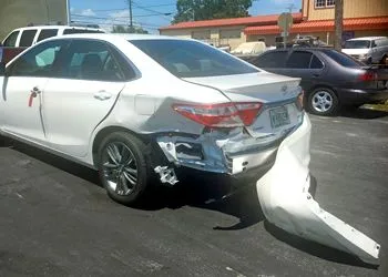 A white car with a damaged bumper is parked in a parking lot.
