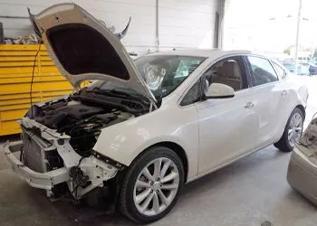 White sedan with front-end damage in a repair shop, hood open and bumper missing