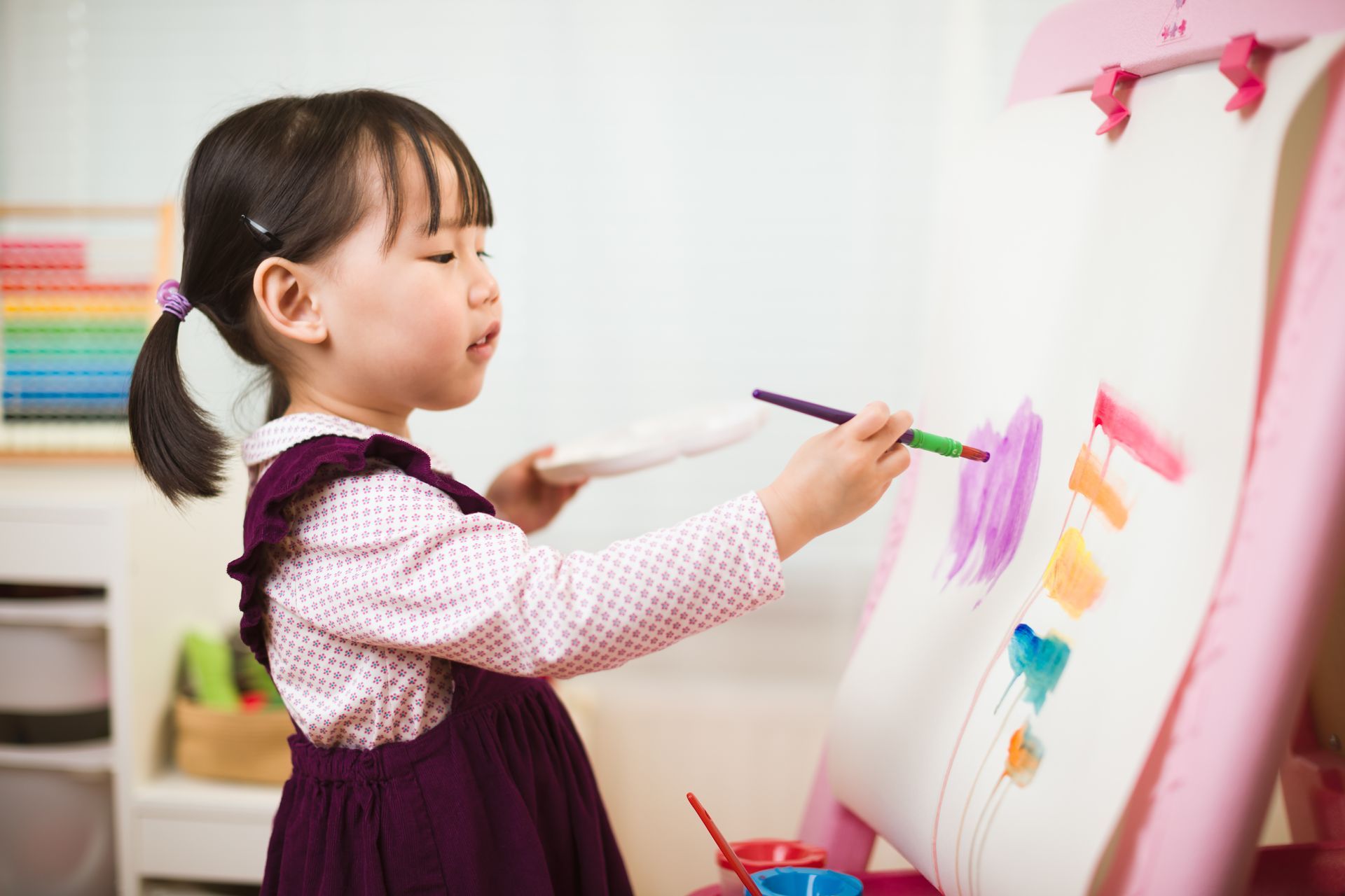 Child painting at an easel with purple paint, holding a palette, inside a room.