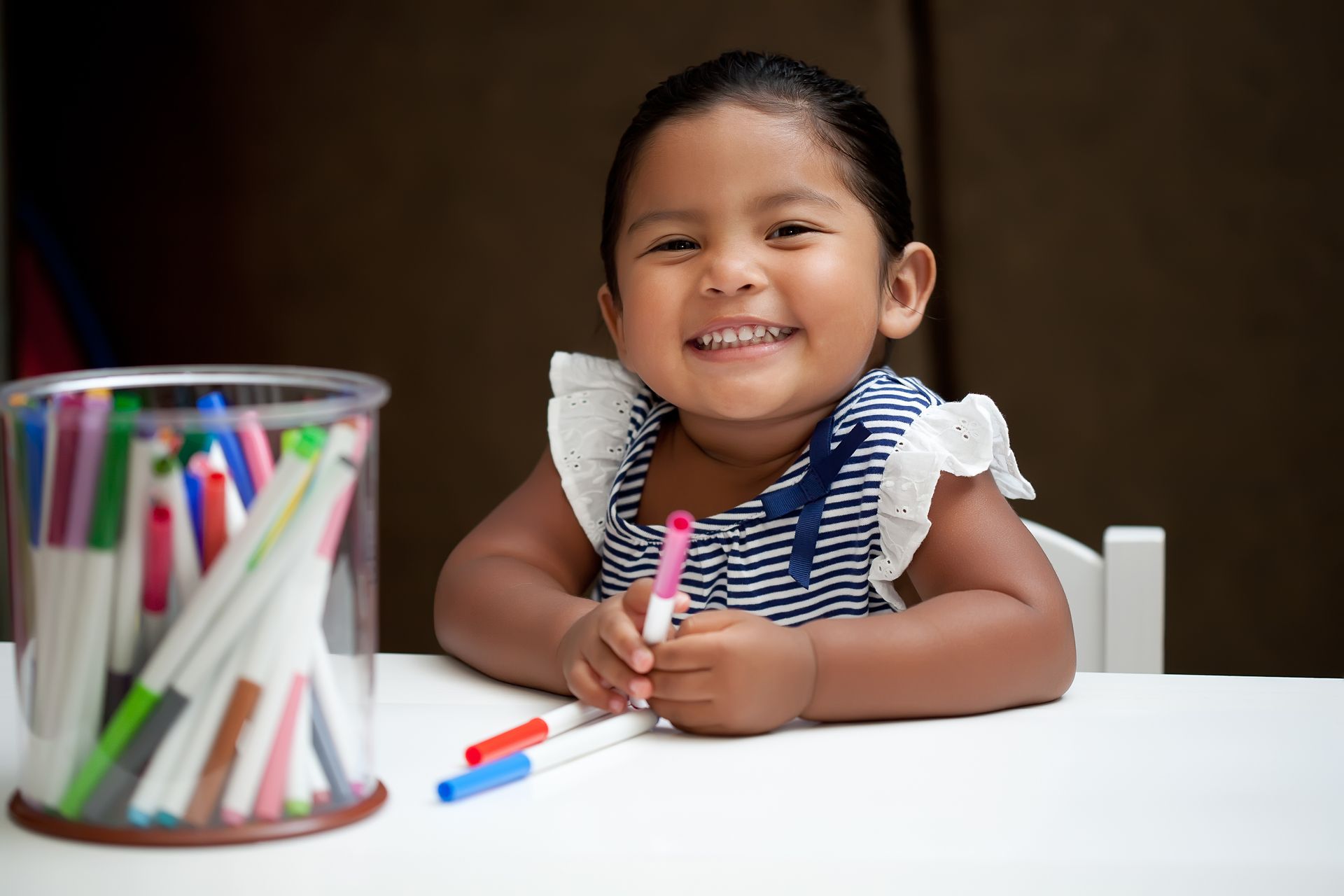 Smiling child at a table, holding a marker. Markers in a jar to the left.