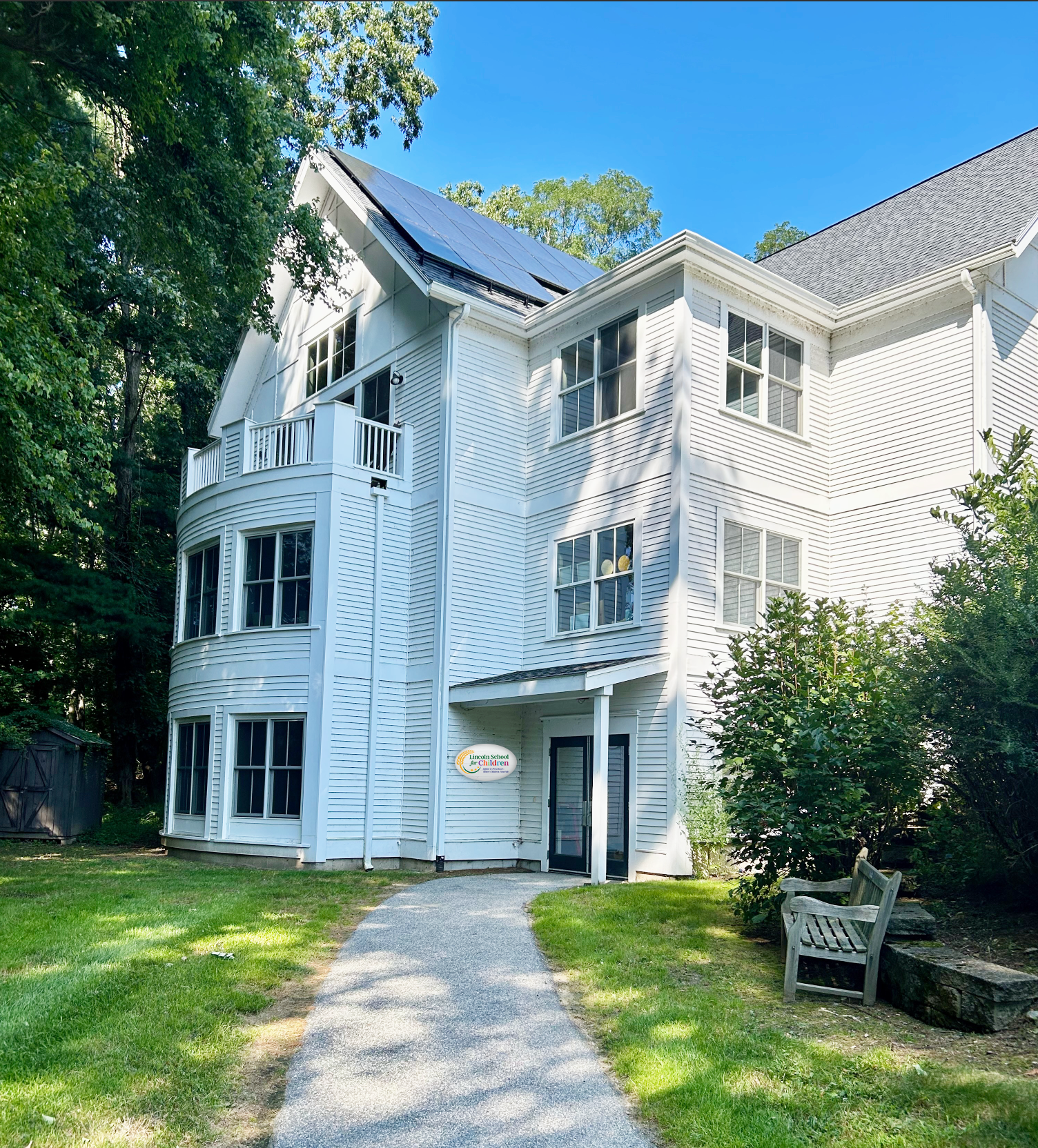White three-story building with solar panels on roof. Gravel path leads to black door. Wooden bench sits on right.