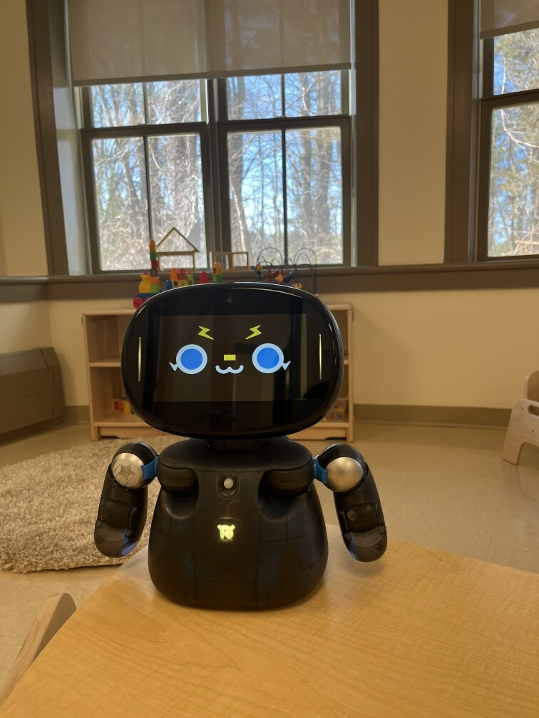 Black robot with animated face, silver arms, sitting in a room with windows and wood shelving.