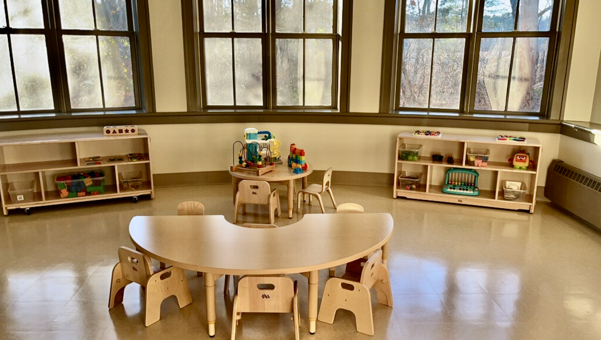 Empty preschool room with tables, chairs, and shelves against large windows.