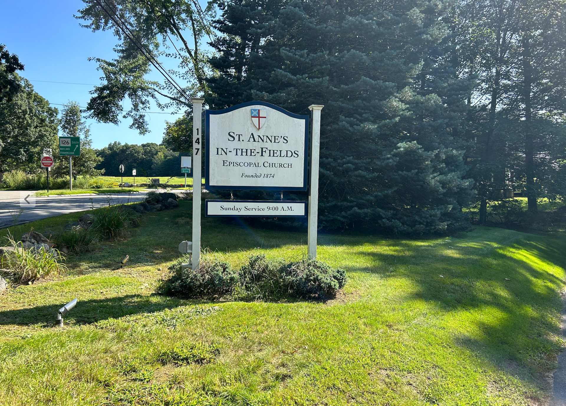 Sign for St. Annes Parish church on green grass, next to a road, under a blue sky.