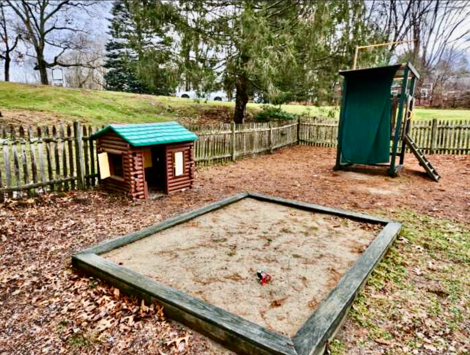 Playground with a sandbox, log cabin playhouse, and wooden tower, surrounded by a fence and trees.