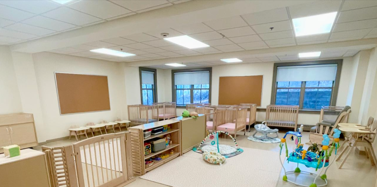 A baby nursery room with cribs, play area, and small tables. Natural light floods the space.