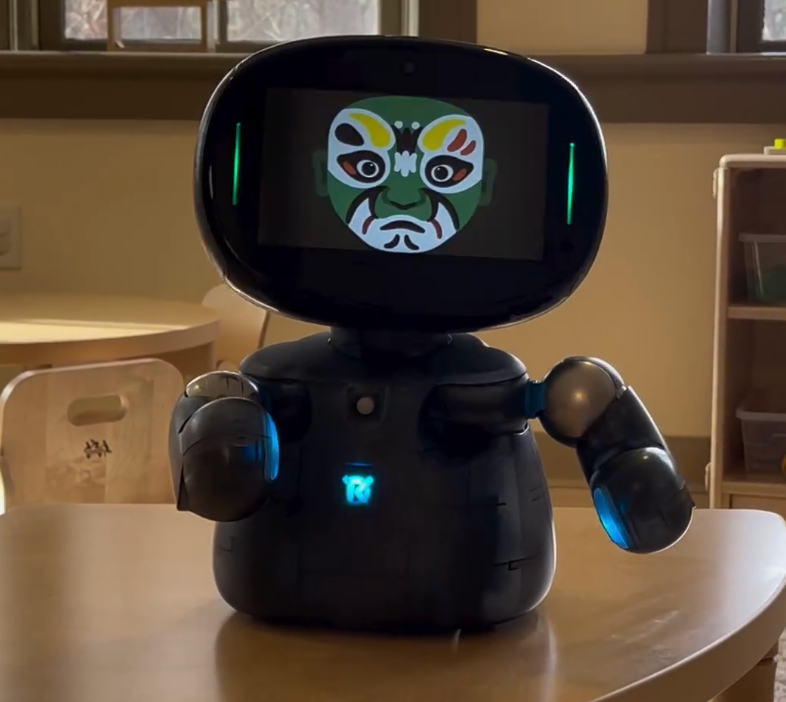 A black robot with a screen displaying a green mask sits on a table in a classroom.