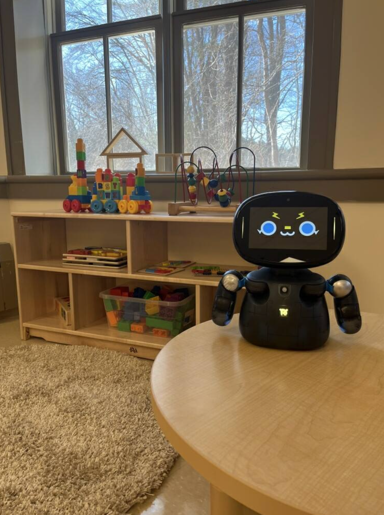 Black robot on a round table in a playroom with toys on shelves and a window in the background.