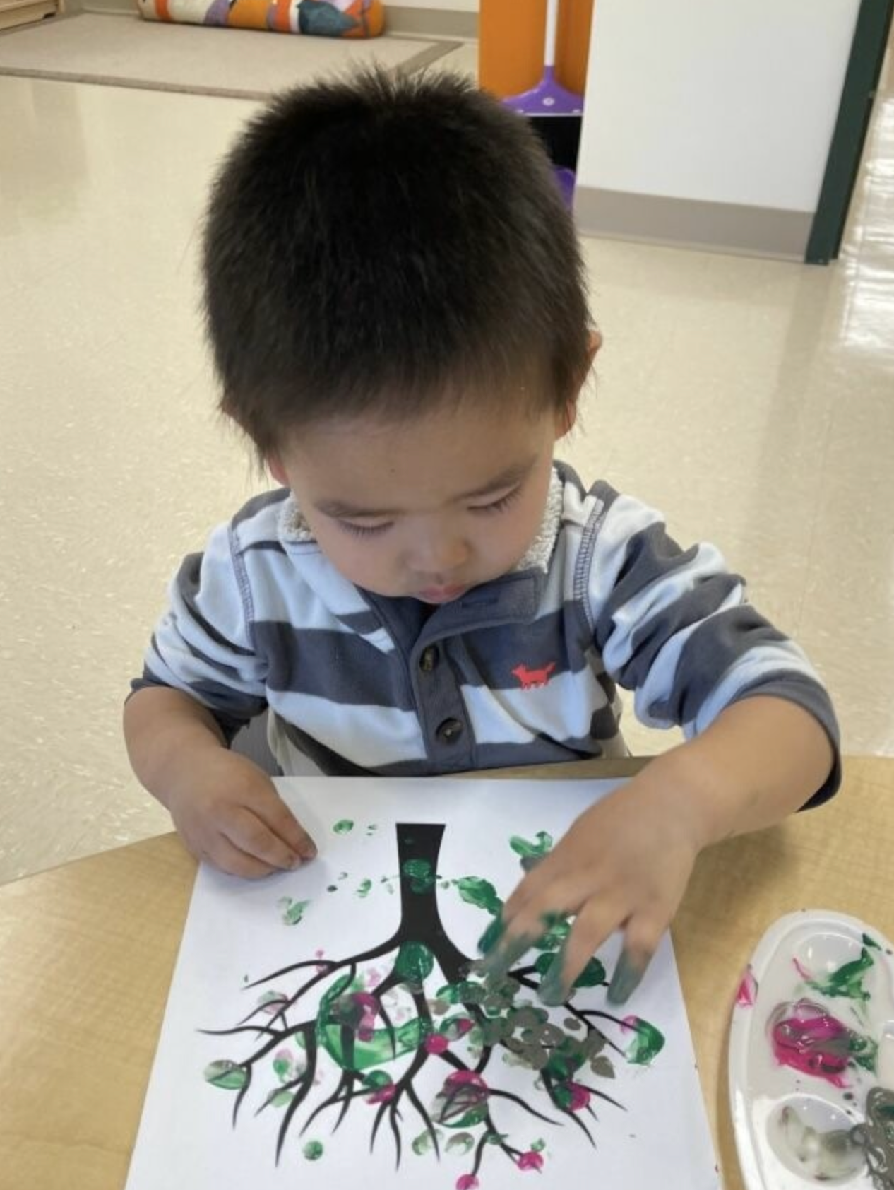 A child painting a tree with green and pink fingerprints on white paper at a table.