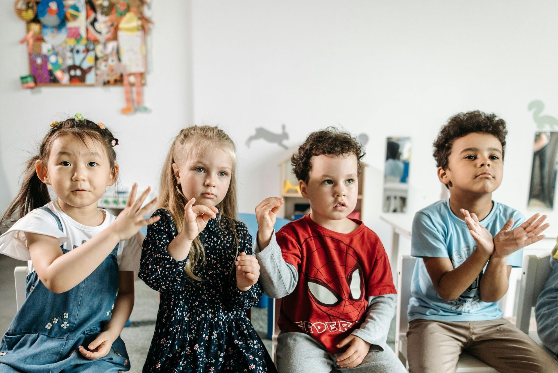 Four children sitting, raising hands, clapping, and looking forward in a classroom.