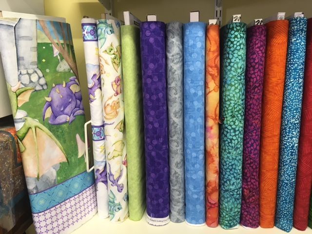 Colorful fabric rolls standing upright on a shelf, including patterned and solid purple, green, blue, and orange bolts