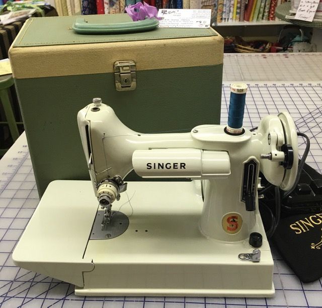 Vintage white Singer sewing machine on a table with thread spool and green storage case behind it Vintage white Singer sewing machine on a table with thread spool and green storage case behind it