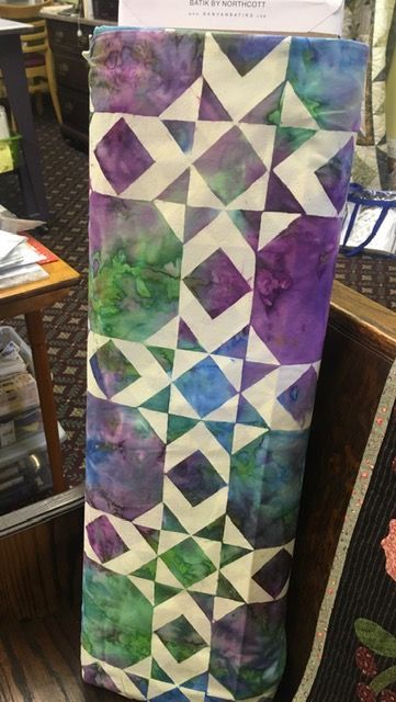 Colorful geometric quilt with purple, green, blue, and white star and triangle patterns hanging vertically