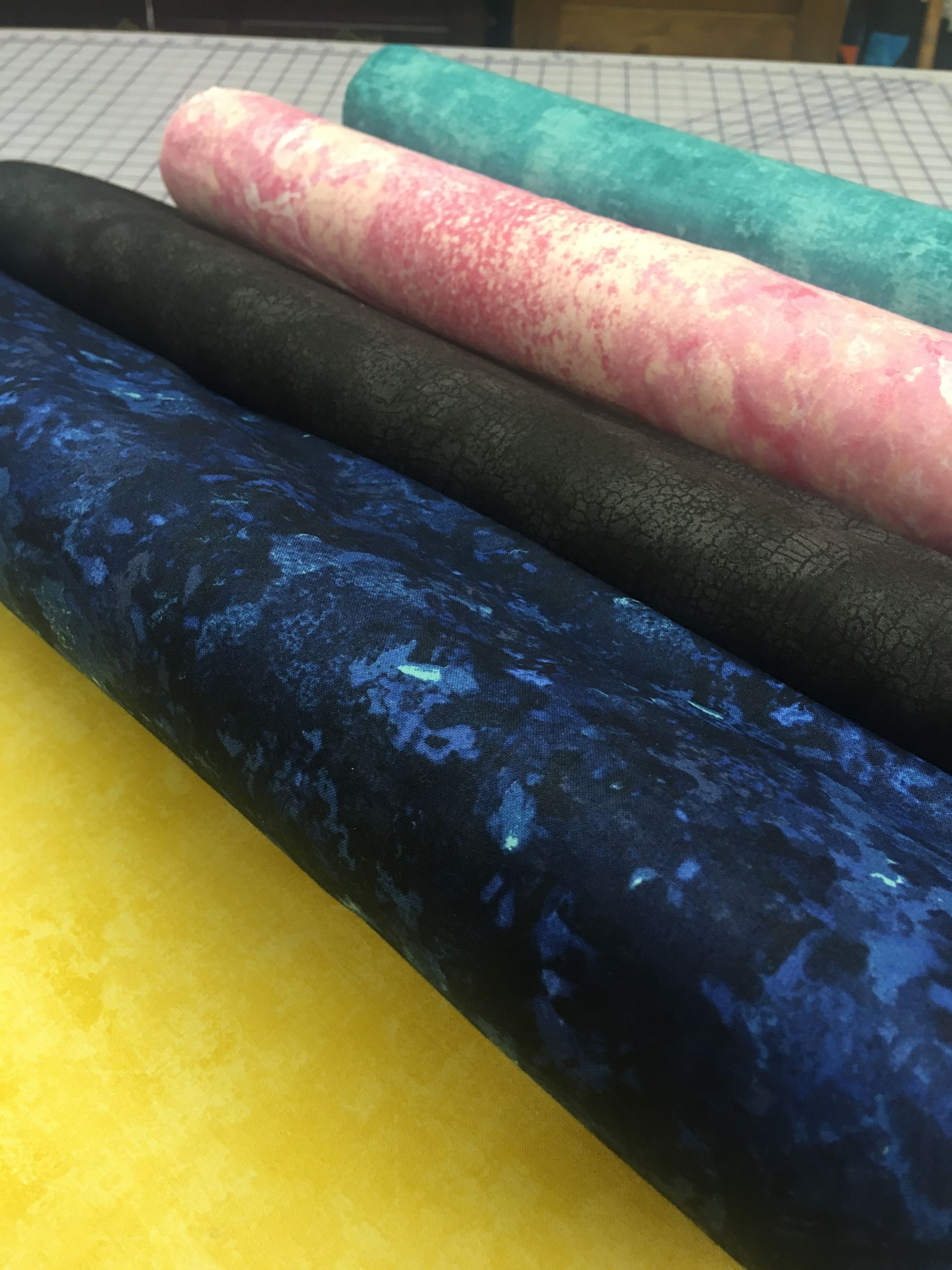 Rolled bolts of colorful fabric stacked diagonally, including yellow, blue, black, pink, and teal.