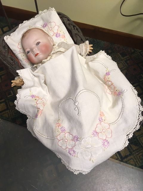 Baby doll lying in a white floral quilt, with a pillow behind its head.