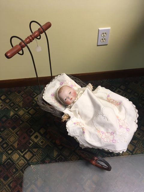 Baby doll in a white dress lying in a basket near a wall outlet and toy handle on patterned carpet.