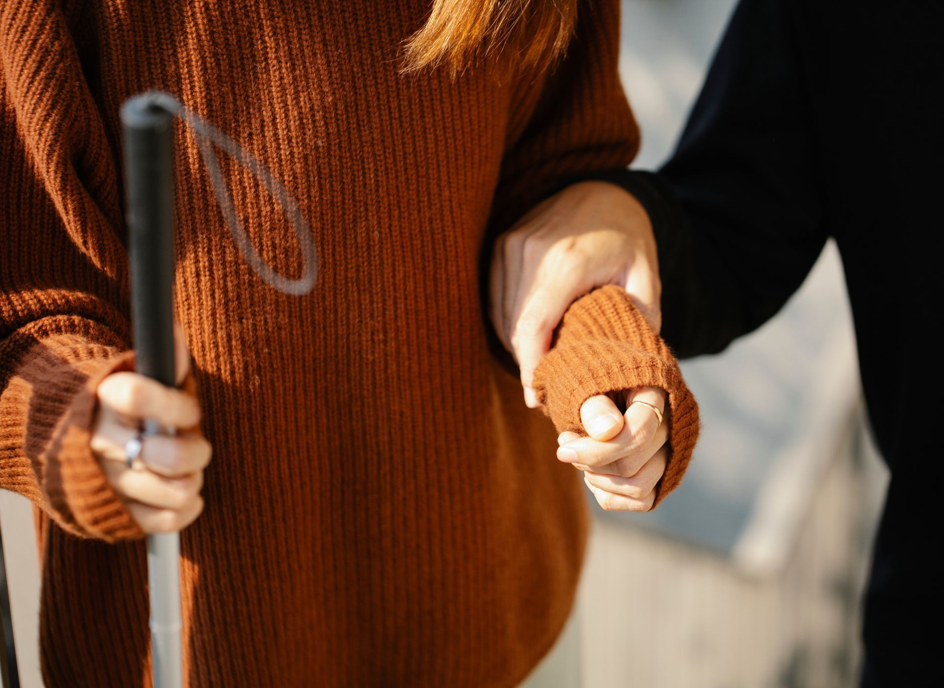 A woman in an orange top and holding a cane is being guided by a lady in a black top. 
