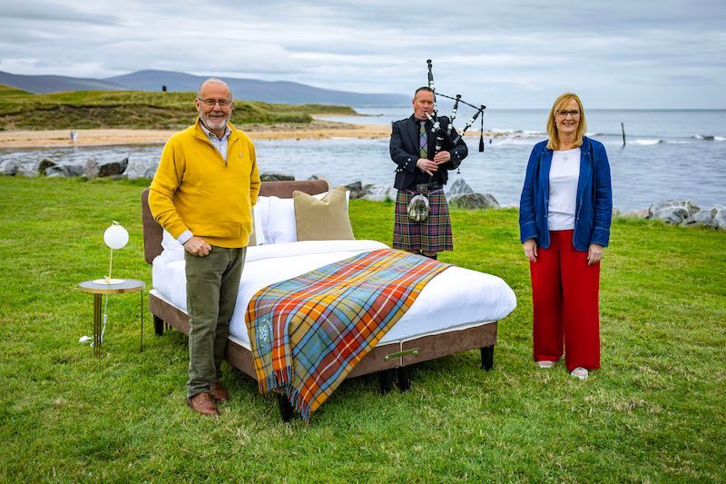 Three people stand on the beach with a bed in front of them. There is a man playing the bagpipes. 