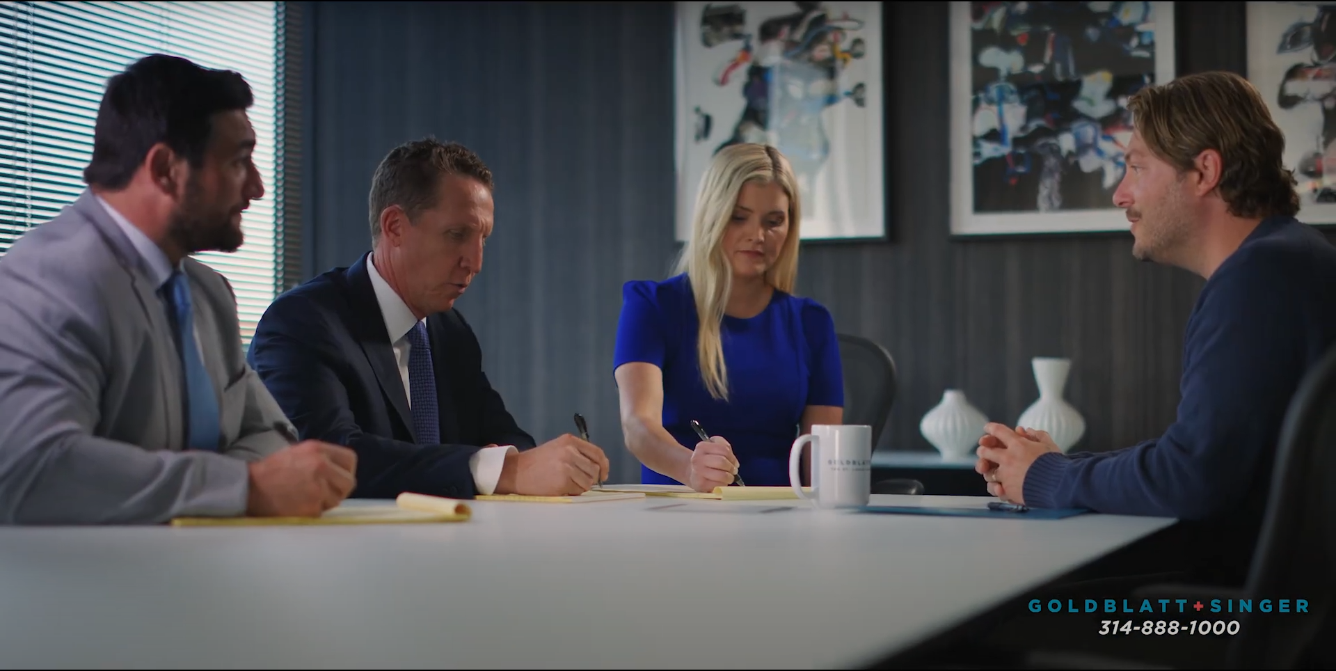 Four people sit at a table in an office. One man speaks to the others who are writing and listening. commercial video lighting
