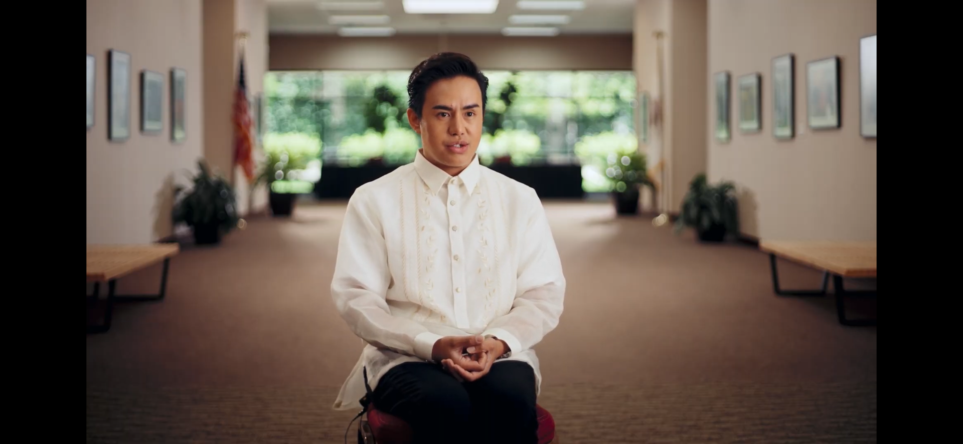 Man in white embroidered shirt sits in a hallway, hands clasped. Brown carpet, neutral walls, and art. corporate video production
