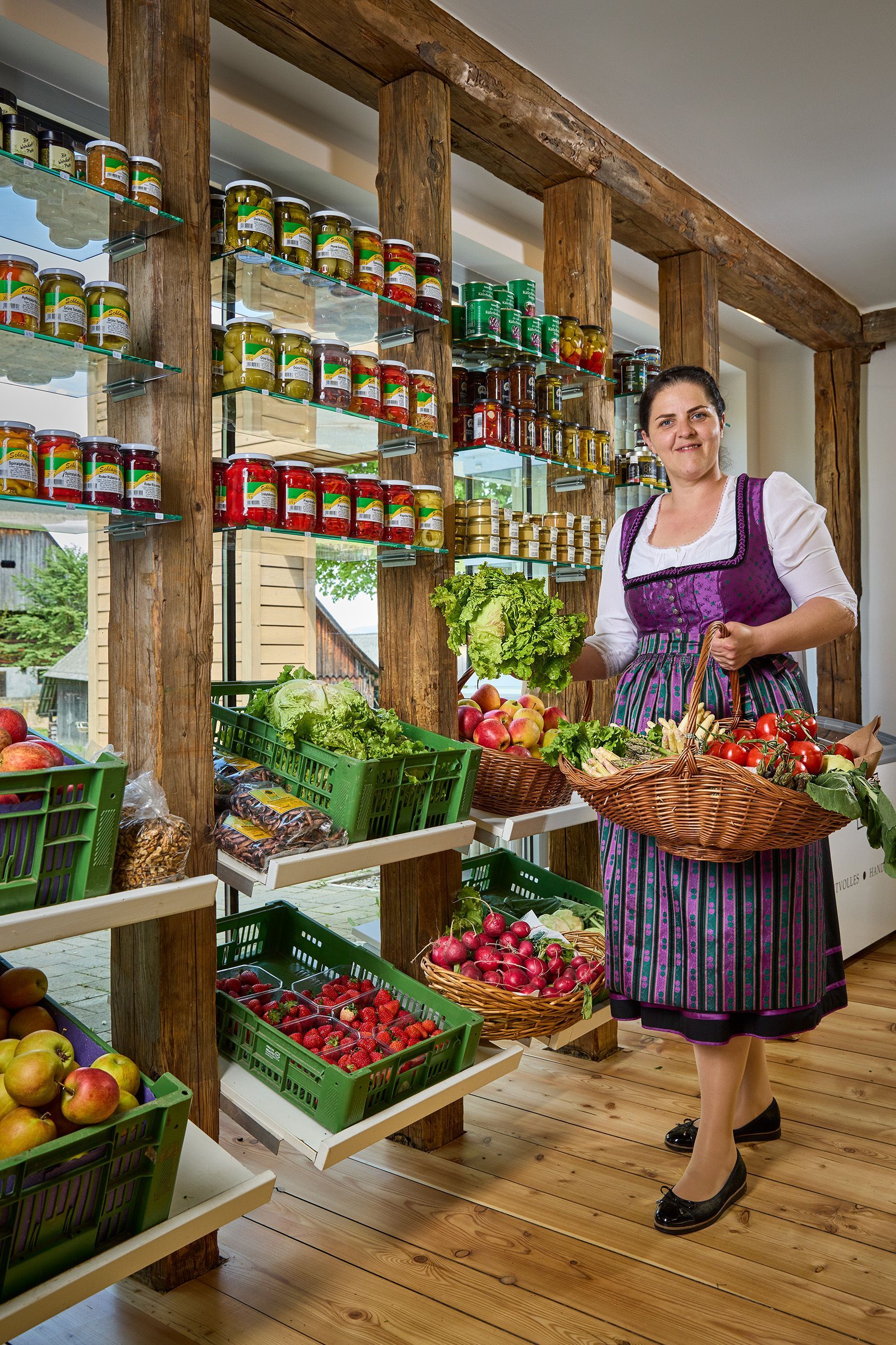 Frau in Tracht in einem Laden, die einen Korb mit Lebensmitteln hält, Regale mit Gläsern, Gemüse und Obst.