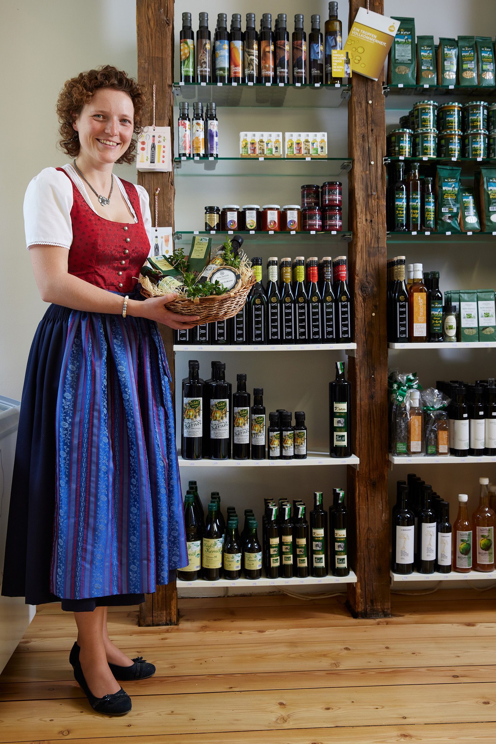 Eine Frau in Tracht hält einen Korb und steht in einem Laden mit Regalen voller abgefüllter Waren.