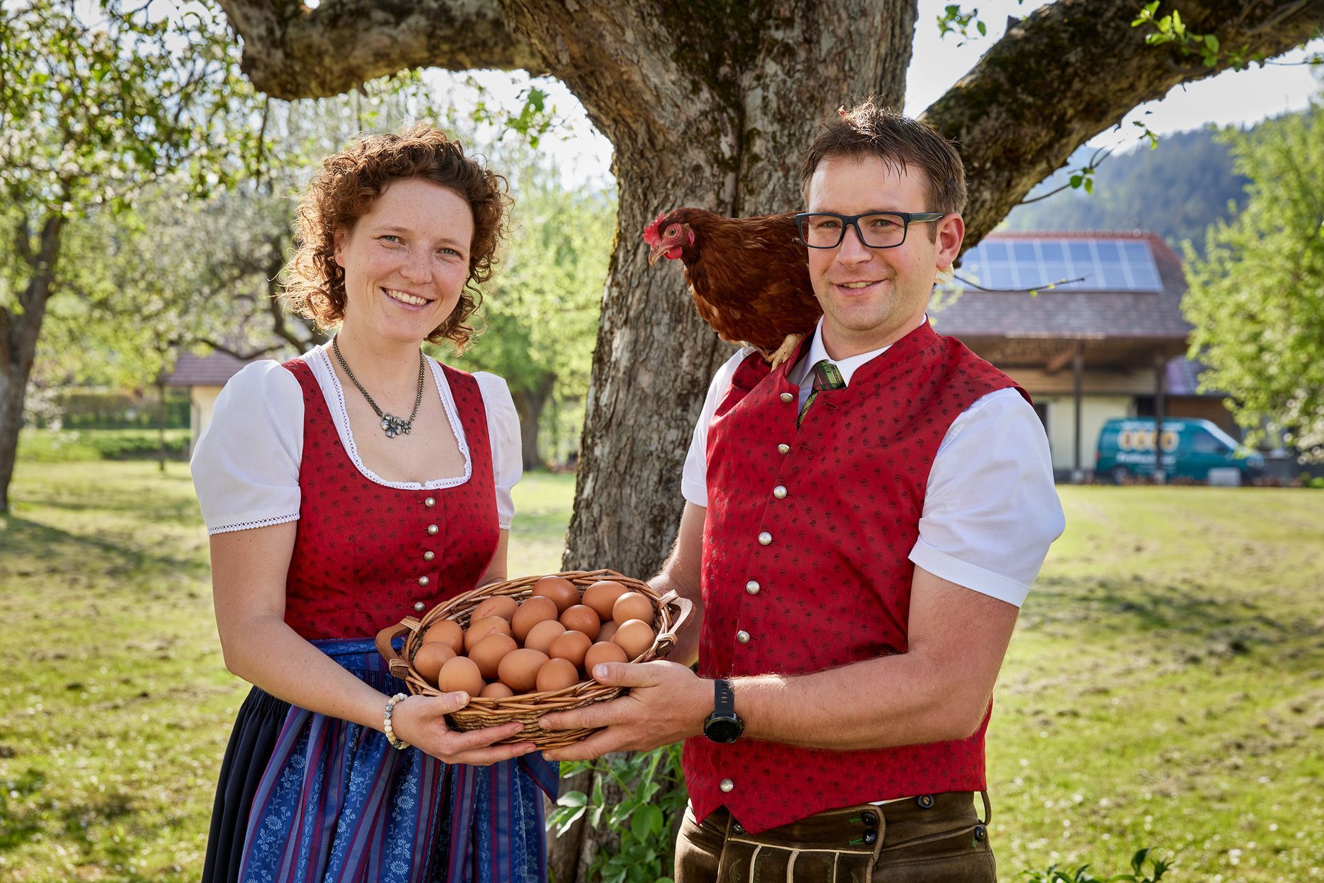 Ein Paar in traditioneller Kleidung im Garten hält einen Korb mit Eiern; auf der Schulter des Mannes sitzt ein Huhn;