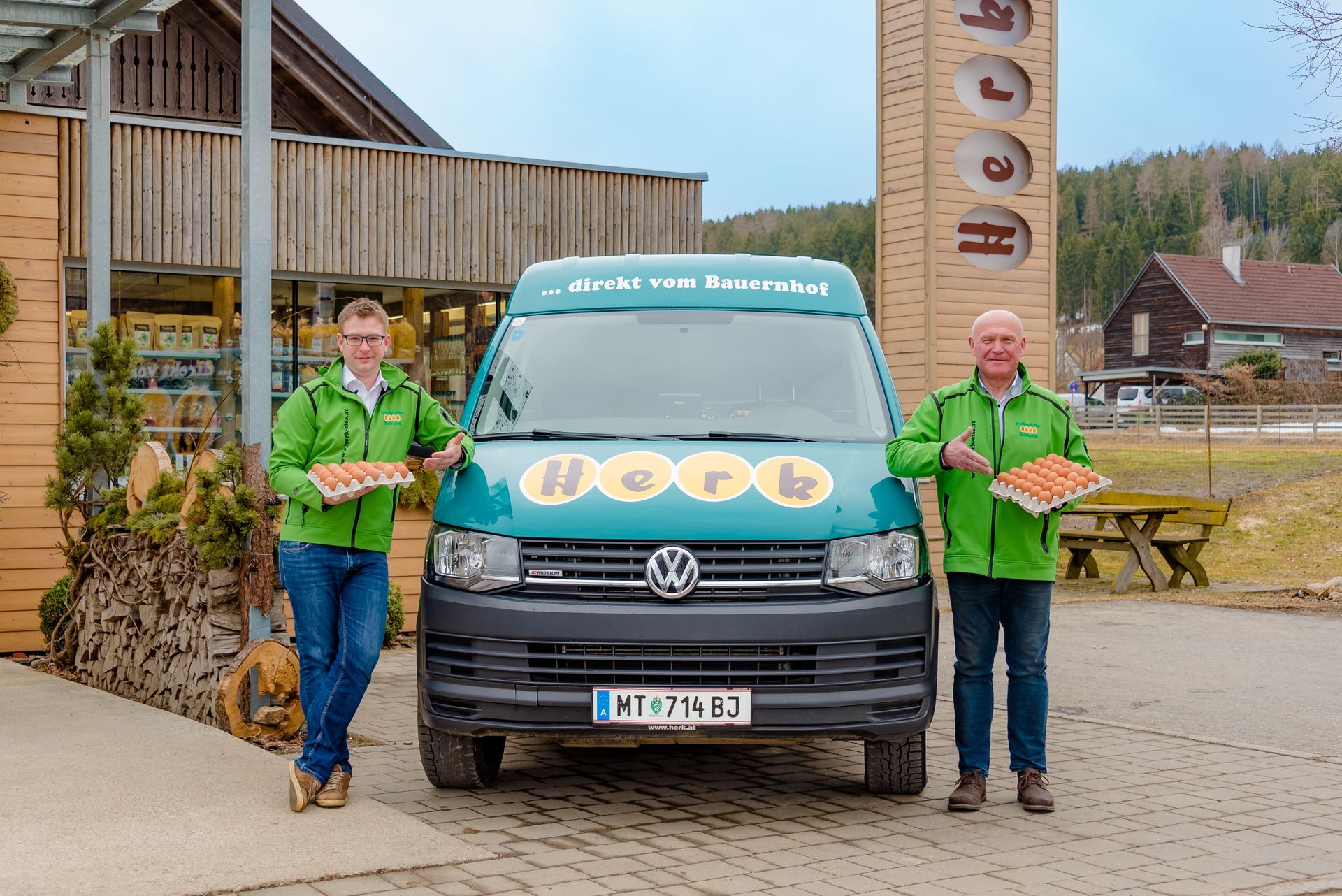 Zwei Personen in grünen Hemden stehen neben einem grünen VW-Bus vor einem rustikalen Gebäude.