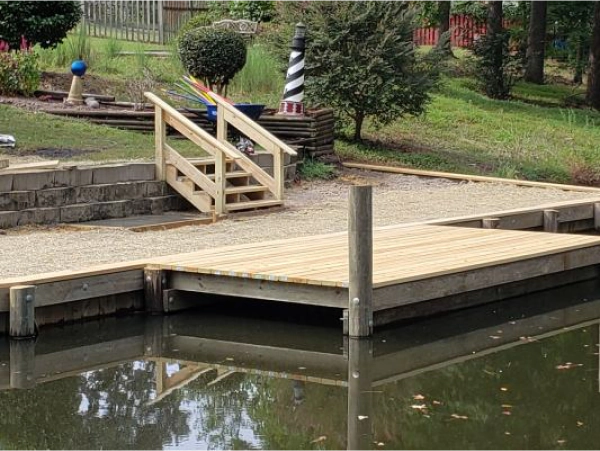 Wooden dock with stairs, steps to a gravel area, in a waterway, with a striped lighthouse in the background.