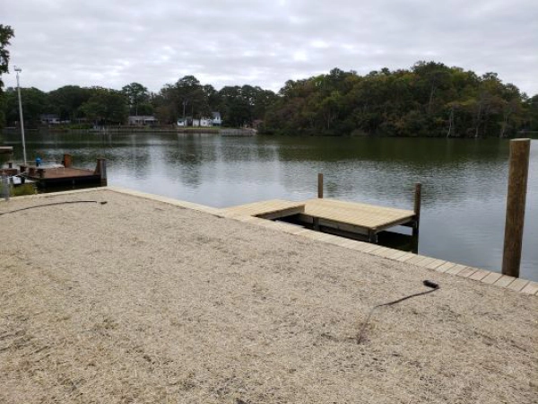 Lakeside view with a gravel patio, dock, and calm water. Trees and cloudy sky in background.